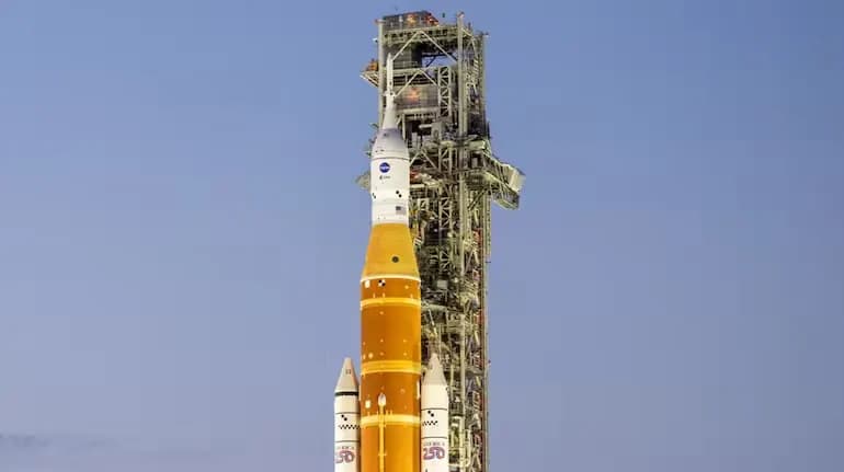NASA Artemis rocket with orange and white sections on launch pad against clear blue sky, tall service tower visible