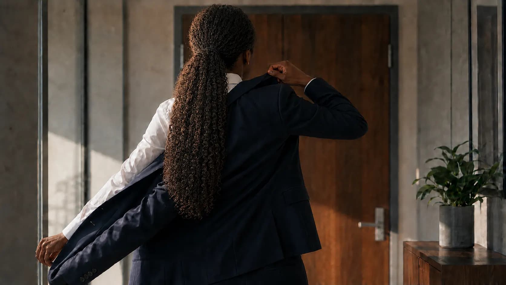 Woman with long curly hair putting on a navy blazer, standing before a wooden door